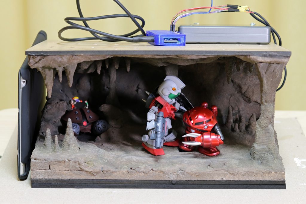 ジオラマの全景。
投稿期限前日まで作業していたのでいろいろ剥き出しですが(汗
MDF穿孔ボード、発泡スチロール、紙粘土が主な材料です。天板はマグネット式で外すことができます。上に乗っている青色の箱がRaspberry Pi Zero WH(LED制御用のワンボードマイコン)、灰色の箱がモバイルバッテリー(電源用)です。
LEDの操作は、離れた場所のパソコンからWi-Fi経由で行います。