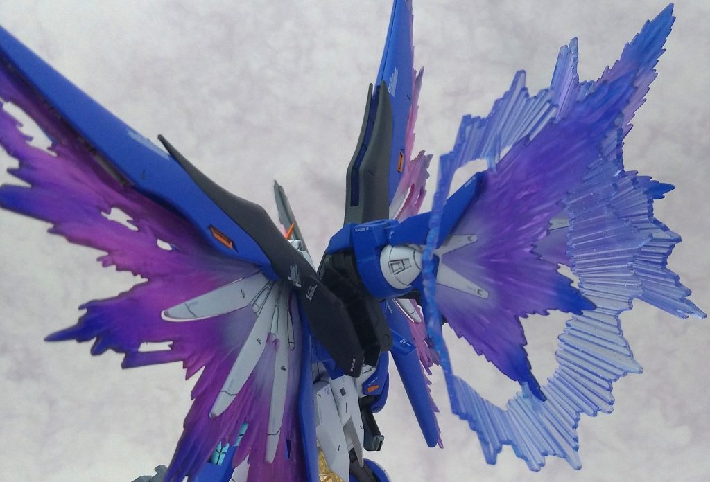 光の翼のカラーパレットについて。
全体にクリアーパープル（G）を吹き付け、根本はクリアーパープルとEXホワイト（G）を混ぜて“強め”のグラデーションを作ります。翼先端はGXディープクリアブルー（C）を適度に乗せ、その後全面にアメジストパープル（C）を吹き、EXクリアー（G）で一層コーティング、という手順で塗装しました。
00スカイの翼も同じ工程ですが、デスティニーの翼はクリアピンクに対しダブルオースカイはほぼ透明なので、最初のクリアーパープルを塗布する際、デスティニーの翼にはしっかりと色を乗せ、00スカイの翼は透明感を僅かに残す程度、リングパーツは全面軽くクリアーパープルを乗せる程度…とパーツによって塗装量を調整しています。
※（G）…ガイアノーツ製品（C）…GSIクレオス製品