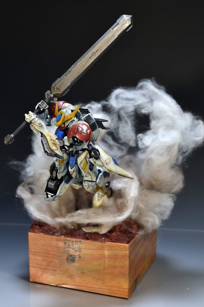 本体の改修箇所としては太ももが上がるように右太もものアーマーの上部をカット。さらに膝も曲がりきるようにふくらはぎの部分をカットしております。
左肩アーマーも独立させ、接着。
 
泥臭い戦闘シーンが魅力の作品だけに、作中イメージを崩さぬよう、全身に軽くダメージ加工と、ウェザリング塗装も施しています。
 
巻き上がった土煙は100均で入手した手芸用の綿を使用。タミヤの透明ソフトプラ棒に両面テープを巻いて固定し、水で溶いた木工ボンドを霧吹きで当てています。