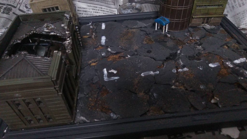 その一方でジオラマの方です。道路は石灰を薄く固め、砕いたものを貼り付けています。割れたところからは土をイメージさせるためバラストをまいています。建築物は鉄道模型用の物を使いました。