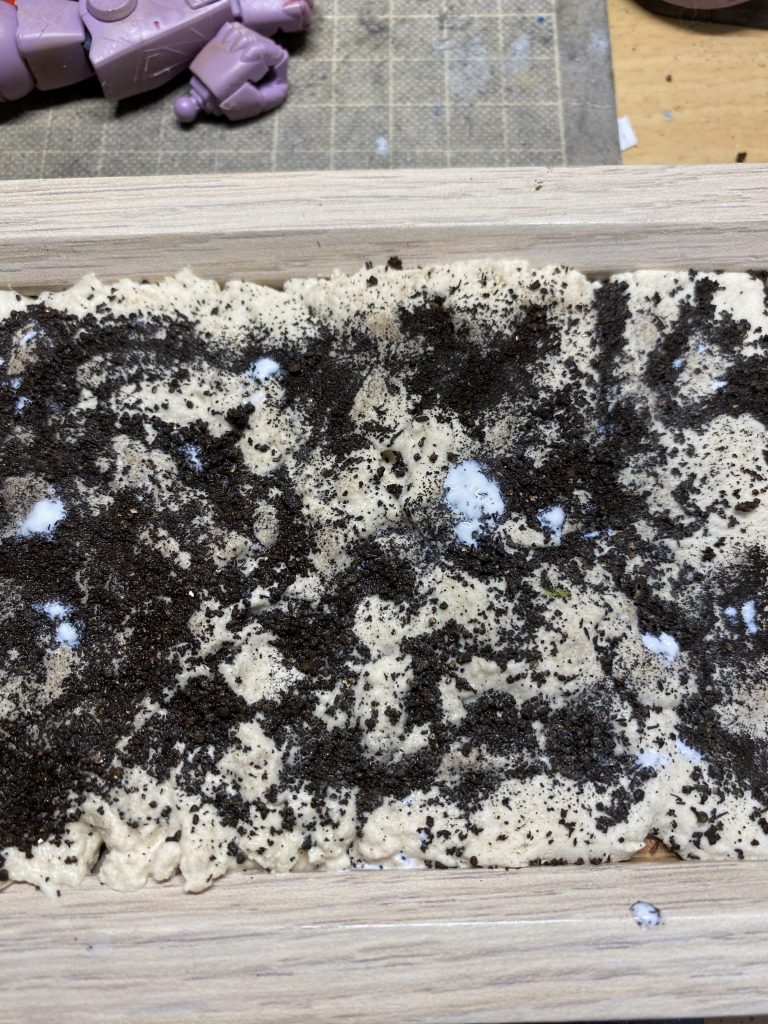 ヴィネットの初期段階、ダイソーの木枠に木分粘土を敷き詰め、乾かないうちにカイロの粉をまき、溶かしたボンドで固めている、