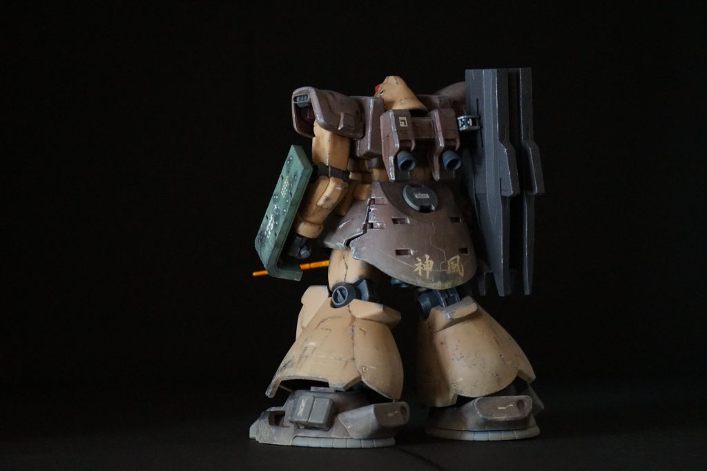 [機体解説]一年戦争末期、本国への帰還が叶わなかったジオンの各勢力は、身を潜め再起を待つ者・各地でゲリラ活動をする者・連邦へ投降する者など様々であった。とあるアフリカの連邦軍駐留基地に、単騎MSが奇襲を仕掛けてきた。周囲に他の敵影はない。「ジムなんか乗ってる奴ぁ、アウト・オブ・眼中！」強気な発言は鳴り響く警告音にかき消され、コックピットは数え切れないアラームの点滅で赤く染まっていた。弾薬は底を突き、残された武器はヒート剣のみ。改修により予備刀身は使えるが、どこまで機体が持つか分からない。次々と連邦のモビルスーツを切り倒すも、次第に機体の損耗が激しくなっていく。応急処置のため60%まで吸気効率の落ちたダストフィルターでは、推力もままならずオーバーヒート寸前であった。「見せつけてやるぜ・・・ジオンの誇りじゃなく、俺のテクニックの凄さをなぁ！！」のちに連邦によって回収された機体の一部に残された「神風」の文字。旧世紀の大戦にまつわる言葉らしく、その意味を知る者は今やほとんどいない。
