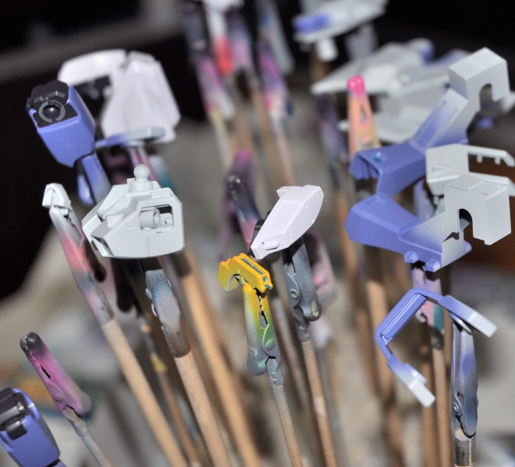 初代AOZのカラーリングについて徒然と今回ヘイズルを作るにあたって一番苦戦したのは色あいです。初代AOZは基本的にダークブルー、紫がかったブルー、ホワイトを基本色に、レッド、イエロー、フレームの紫っぽいダークグレーが使われており、T3カラーでもテスト機カラーでも実戦配備カラーでも、その割合が変わるだけで基本的に使われている色は変わりません。なのでAOZ系のキットを作っていく予定で、かつ色合いを統一したいという想定であれば、可能な限り調色しないで出来合いの塗料を使用する、あるいはごく簡単に調色できるようにする必要があります。上述したような単純な塗料選択はそういった理由からであり、またAOZ特有のカラーにマッチする塗料が多く発売されたために実現可能になったと言えます。…が、実は初代AOZのイラストというのは、「同じ色のはずなんだけどイラストによって全然色合いが違う」という大きなが罠があります(笑)なので、あっちのイラストにあわせて、これだ!という塗料を見つけても、いざテストしてみるとなーんか違う色に見えるという事態になります。ダークブルーで言えば、割と青みを感じイラストもあれば、真っ黒に近い時もあるしブルーで言えば、割と赤みが強い「紫」感が強い明るいパープルブルーの時もあれば、どちらかというと青みが強く、くすんだ感じで、ブルーグレーに近い時もあれし、コバルトブルーのような青に見えるイラストもある、という具合ですなので、ばっちりイラストの色合いに合わせていく、ということはAOZ系キットを共通の色で塗ろうと企てるかぎり不可能であり、求められるのは、それらの平均値のイメージに合致する色を探し出すことだ、と言えます。・・・作り始める前にこの結論に達するまであーでもないこーでもないと悩み、たぶん半年から下手すると1年ぐらい行ったり来たりしてたというAOZってメンドくせーな!(誉め言葉)というお話でした