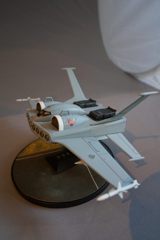 左のエアインテークにはWW2の爆撃機的な「ノーズアート」を入れてみました。エアインテーク上部にはモビルスーツ用のフックを取り付けました。また、以前から思っていたのですが、高速飛行物体に載るのにいくらMSとはいえ、浅い凹みに仁王立ちでは、すぐに振り落とされちゃうと思うので、MS搭載アタッチメント的なものをジャンクパーツで適当に設置しました。スノボの板にブーツを固定するバインディング(スロットイン)的な感じです。