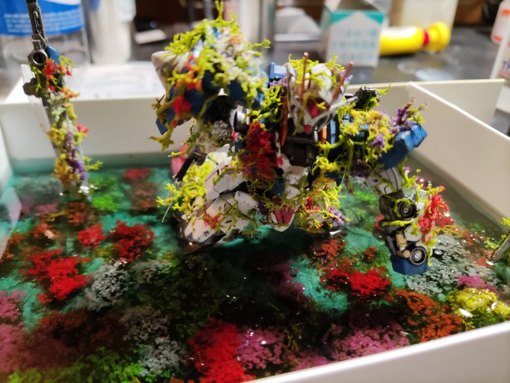 一方、ジオラマの土台部分となっている「湖に沈む花畑」部分は百均のフォトスタンドに紙粘土でベースを作り、リューターで削って石畳を成形。アクリル絵の具で色付けし、水溶きボンドでコーティング。その上に「草むらと花セット」を適当な大きさに刻んでセンスよく??配置していきました。最後にGNソード2×2本とダブルオーガンダムを配置してレジンが漏れ出さないようにプラ板と木工ボンドで養生し「日新クリスタルレジン300ｇ」を薄い緑に着色して二度に分けて流し込みました。今の時期（7月上旬）で半日ほどで硬化、二度目は薄いブルーに着色して流し、同じく半日で硬化しました。最後にプラ板を剥がし、レジンのバリを取ったら完成です。ジオラマもレジンも初めて扱うので正直不安でしたが思ったより大きな失敗はありませんでした。