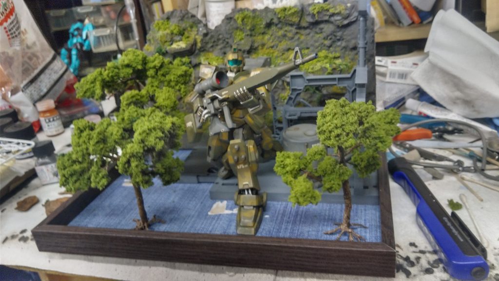 樹木や基地部分、岩壁を置いてレイアウト模索中…。最初は右手前に樹木を配置しようと思いましたが兵士達が見えなくなってしまうので後にレイアウト変更しています。基地部分はバンダイのシステムベースを流用しています。