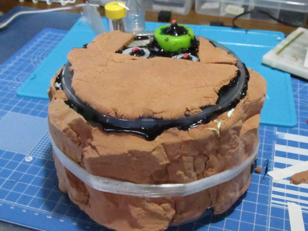 これに軽量紙粘土で作った岩盤を巻きつけてグルーガンなどで接着。隙間も粘土で埋めていきます。片膝を立てたポーズにするための出っ張りとしてペットボトルキャップを使用。もはや完全に小学生の夏休み工作です。