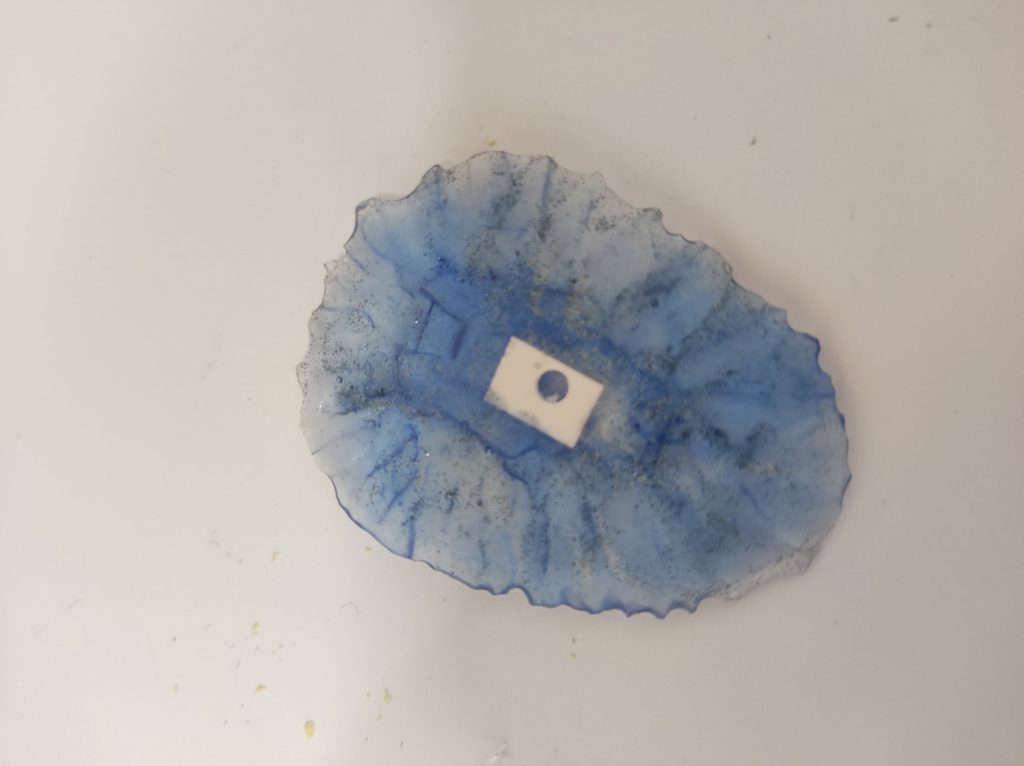 そこに、100均の透明のレジン流して、ユニコーンパウダーを一振り、ブルーのレジンを上から流して、シールド基部に3mm穴開けやすくするため、と、レジンの節約のためプラ版を埋めて固めました。イメージとしてはプラフスキーパーティクルクリアなんですが、中に入れるパウダーが黒系だったので全然違いますね