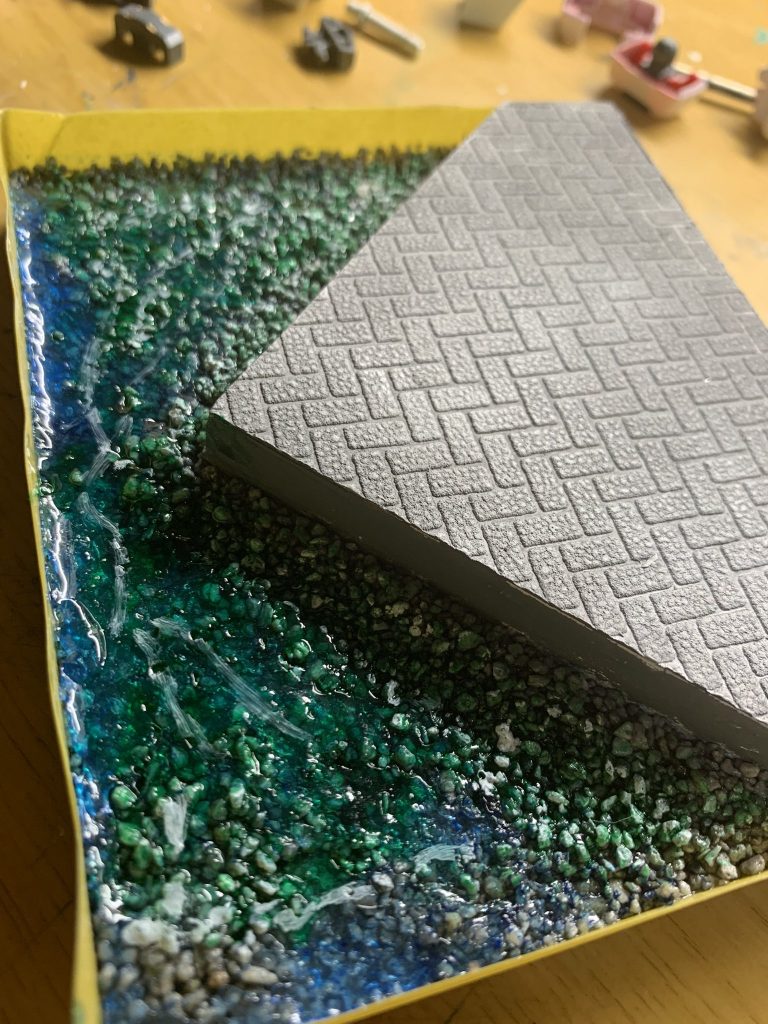 鉄道模型用かなにかの砂利を水溶きボンドの海に撒く。乾き次第青い透明レジンを一層……気泡抜きに1日置いた後にUVライトで固め、白やクリアグリーンで模様をつけてまた一層……これを繰り返して……