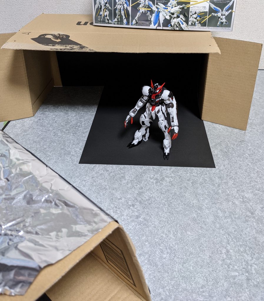 撮影風景。ダンボールに100均黒紙背景です。箱が浮くのでバエルでおさえました。