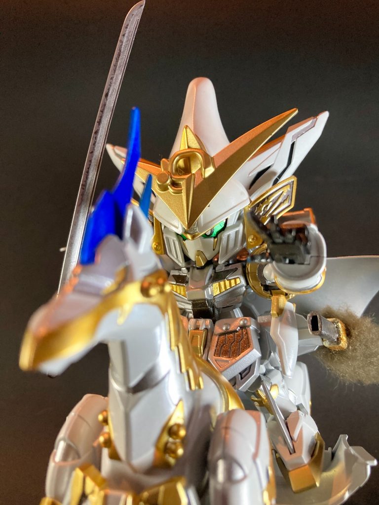 〜〜〜〜〜〜〜〜〜〜〜SDWヒーローズ・オリジナル英傑を製作しました。ヒーローズ本編の第4話、信長ガンダムエピオンがムシャワールド内で敵対する大名として「謙信、信玄、元就、勝頼」と言及するシーンがありました。その後しばらく第7話の本能寺ホテル宿泊の頃には天下統一の目前まで進んでいたので、彼ら諸大名が実際に劇中に登場する事はありませんでしたが、この「各地に戦国大名が割拠するムシャワールド」という世界観が示された事から着想を得て、今回上杉謙信モチーフのキャラクターを作らせて頂きました。演者MSはマスターガンダム、ベースキットは三国創傑伝の典韋マスターガンダムです。カラーリングを上杉謙信イメージの純白へガラリと印象変える事で、元の典韋とは全く別の人物な雰囲気に仕上げています。造形はマスターガンダムの特徴的形状を基本に残しながら、SD戦国伝・武神降臨編の上杉謙信頑駄無のキットから戦国武将らしいパーツを随所に仕込みました。謙信所用と伝来の日月前立を額に、ガンダムヘブンズソードの頭部も少し意識した長烏帽子形兜はエポパテを盛って削って整形しました。謙信頑駄無の肩鎧はマスターガンダムの背部バインダーに見立てる形で装着し(騎乗時は母衣のような膨らみが感じられて良い感じになりました)、腰部フロントアーマーの左右にも甲冑パーツを利用しています。