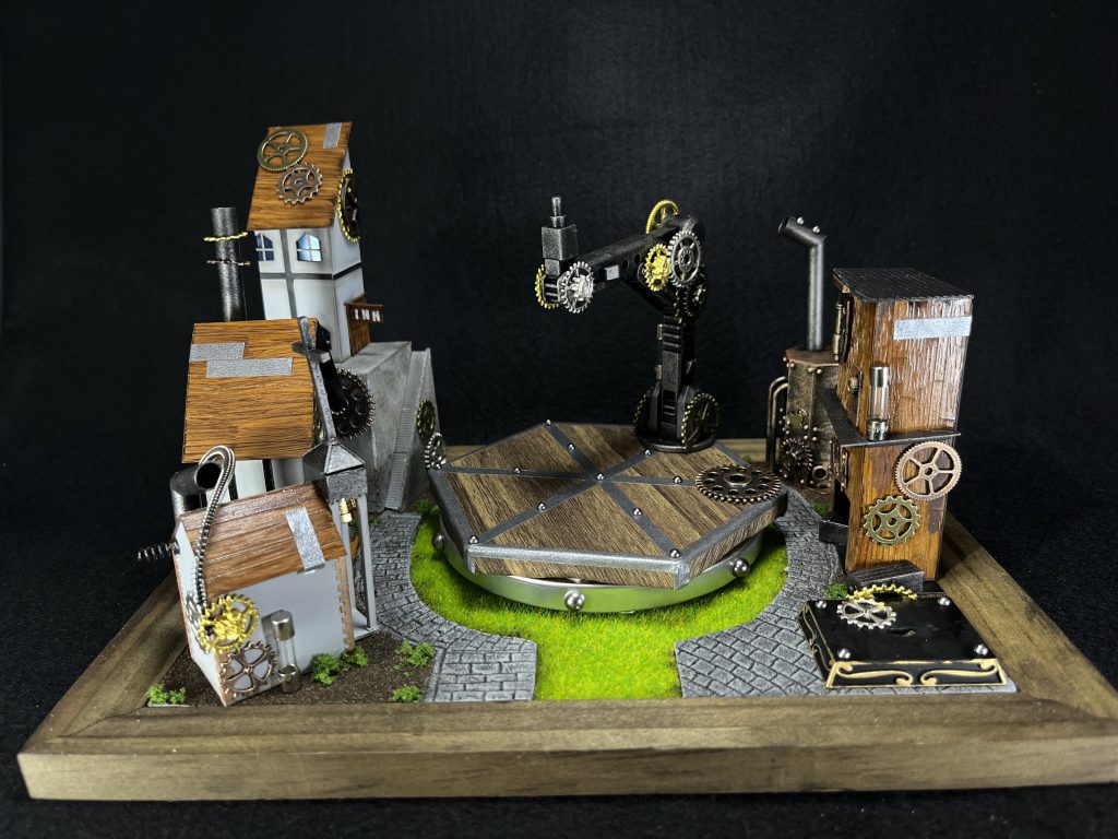 今回のジオラマは、スチームパンクな街の守り神的な存在として街の広場に鎮座している状態をイメージして作成しました。アクションベースをジオラマの一部にするため、木目シートを貼ったり、歯車つけたりで雰囲気を出しました。周りの建物はプラ板の箱組みにて作成し、新しい技術として木目塗装に挑戦しました。タガネやケガキ針で木目をつけた後、薄めのベージュみたいな色をスプレーし、ウェザリングカラーのステインブラウンを木目方向に筆塗り、仕上げにクリアオレンジをスプレーしました。屋根や右側の建物の壁に使用しましたが、結構ちゃんと木の感じ出たと思います。