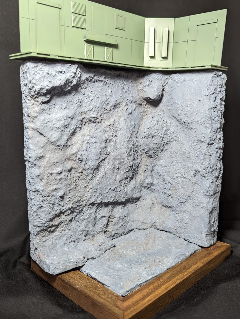 背景はフォトフレームに、紙粘土を引き詰め形をそれっぽく作り、岩場はスタイロフォーム立ててカッターとハンダゴテでそれらしく形を作りました。大きなでっぱりは、スタイロフォームの切れ端をボンドで着けています。大まかな形ができたら、トイレットペーパーを少量の水と木工用ボンドで溶いて、それを塗って凹凸を強調し、乾いたらセメントを塗っています。塗装は画材絵の具でそれらしくしています。全て100均で購入しています。セメントはダイソー、それ以外はセリアのモノです。上の装甲的な部分はプラ板でテキトーに作っています。色はガンダムカラーの緑です。量産型ザクの塗料はジオンには大量に余っているからだと設定しての着色です(笑)