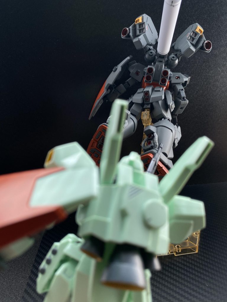 「速え!まるでバッタだ!」 戦闘機動、凄まじいGに耐えながら、ジェガン二番機のパイロット、アラン・ガヴィルは吠える。「ロックオンさせてくれないっ」 一番機のパイロット、セルマ・コトネンの声。その声がはっきりと聞こえるのは、ミノフスキー粒子が散布されていないからだ。連携していきましょう。セルマの声に対し、フォーメーションを組む事で応える。相手はカスタムされているとはいえ、ジムなのだ。「こっちはジェガンだぞ、何で付いてくのが精一杯なんだよ!」 瞬きすら許されないスピードで大小様々なデブリを躱しながら、吠える。「落ち着いて、アラン。宙域図(マップ)を」 もう見てるよ。そう言いながら、相手の予想進行ルートを掲示する。それに対し、セルマは自分と違う予想進行ルートを表示する。巨大な残骸(おそらくマゼラン級だろう)の脇をかすめるルート。見ると、相手のスラスター光が巨大な残骸の方に向かうのが見える。さすがセルマだ。先からの断続的な射撃で、残骸の方に誘導していたらしい。「残骸を抜けるタイミングで挟撃しましょう、左右!」 いや、上下だ。天地そのままの状態で艦橋の手前から折れてしまっているマゼラン級の後部から侵入するルート、初めは左右から回り込む、それぞれのタイミングで上下方向に急転回。ジェガンの機動性能と頑強なフレームがそれを可能にする。「セルマは上、俺は下から」 機体を捻るように旋回させ、マゼランの下方へ、セルマも艦橋を超えるように動いているはずだ。あの背中のブースター、ジムよりジェガンにつけたほうがカッコいいよな。そんなことを思いながら残骸を回り込む。このタイミングなら、相手が残骸を抜けたタイミングで挟撃できるだろう。