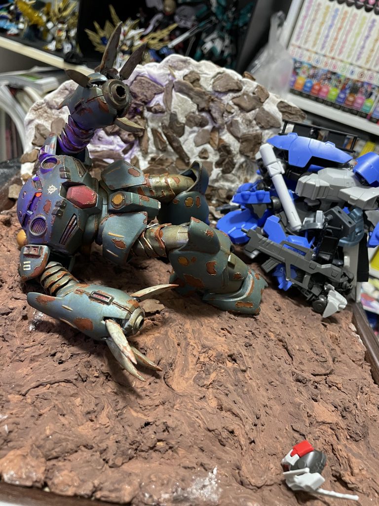 ジオラマ制作途中。最初は水から上がってきたところみたいのにしたかったのですが、水の表現はめんどくさそうだったので、とりあえず練習として土と岩をやってみました。土はコルッキーという土の表現ができる粘土をダイソーの写真立て敷き詰めただけです。何もしなくても本当にそのままでちゃんと地面になるので初心者にはちょうど良いと思います。岩はダイソーの紙粘土にダイソーのバークチップという木のかけらみたいのを貼り付けて、ダークグレーで塗装してからホワイトでドライブラシをすると完成です。ジャンク山はテキトーです。ジャンクのサビは、流石にズゴッグEほどの手間をかける気力がなかったので手抜きです！今回初めてサビ表現やジオラマをやってみましたが、今はすごく便利な道具があるのでど素人でもそれっぽいものが簡単にできたりしてめっちゃ楽しかったです！！！でもかなり場所取るのでもうやらないかも！置く場所がない！！！