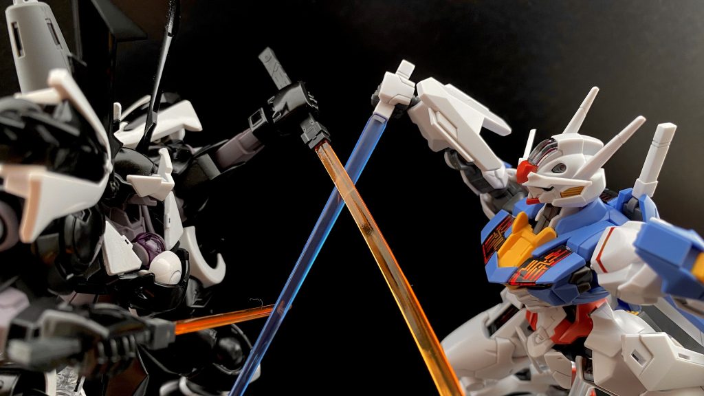 　ぶつかり合うビームサーベルのスパークを浴び光るエアリアルの顔を見据え、吠えるグラハム。「私のものになれ！ガンダムッ!!…もはや愛を超え、憎しみを超越し、宿命となった！」　スレッタもまた、サーベルの光に照らされる黒いハインドリーの頭部に向かって声を張り上げる。「わかりません、あなたが何を言っているのか全っ然わかりません！」「でも、これだけは言えます。ガンダムはっ、エアリアルはっ、あなたのものじゃありません！」　互いのサーベルが弾かれる。頭の上に弾かれたサーベル、向かい合う二機のMSはそのままサーベルを大上段に構える。あとは、振り下ろすのみ。「私がっ！」　スレッタが叫ぶ。「私たちがっ！」「(株式会社)ガンダムですっ!!」「なッ、なにッ！」　一瞬だった。一瞬振り下ろすのを遅れた黒いハインドリーの腕の隙間を、エアリアルのサーベルが、一閃する。　…決闘終了の合図が鳴り響く中、ブレードアンテナを根本から断ち切られたスサノオウのコクピットの中で、どこか満足そうなグラハムはつぶやく。「見事だ。ガンダム…いや、スレッタ･マーキュリー」　心の底からの声を聞いたような気がした。その瞬間、レバーを握る手の動きが止まったのだ。