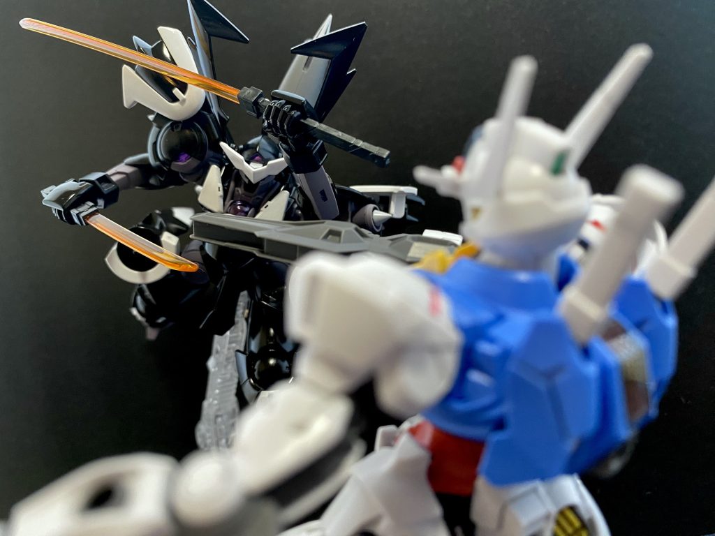 　その駆動性能、推力で組み敷かれた状態から一旦脱出したエアリアルだったが、黒いハインドリーから距離を取ることが出来ずにいた。ライフルや、ガンビットでの攻撃をしようと距離を取るが、黒いハインドリーの加速性能は高く、すぐに懐に入られてしまうのだ。　スレッタが焦っているのが、中継を観ているチュチュ達にも分かる。「オ、オイ、だんだん押し込まれてね」「ハインドリーってのは、そんなに性能が良いのかよっ」「連携戦闘重視の機体だから、単体での性能はそれほどでもないはず…」　そんなチュチュ達の会話など、コクピットのグラハムには聞こえる筈は無い。しかし、今まさにエアリアルを切り裂かんとビームサーベルを振り上げるスサノオウのコクピットの中で、グラハムは裂帛の気合いと共に叫ぶ。「どれほどの性能差があろうとも！」「今日の私はっ！」「阿修羅すら凌駕する存在だッ!!」　グラハムの振り下ろした渾身の一撃は、エアリアルのビームライフルを中程から両断する。エアリアルの回避行動の方が早かったのだ。しかし、スレッタの動揺は大きくなるばかりだ。矢継ぎ早に繰り出される黒いハインドリーの攻撃に動揺を隠せないスレッタに、「おちつきなさい、スレッタ」　冷静な、ミオリネの声が届く。「いい、落ち着いて思い返して、相手はワケの分からない事を言いながら真っ直ぐ突っ込んで来るだけ、操縦技術も、MSの性能も、アンタの方が上！」「み、ミオリネさん…」「それに、アンタまだ前に進んでないでしょ。…進めば…、でしょう」