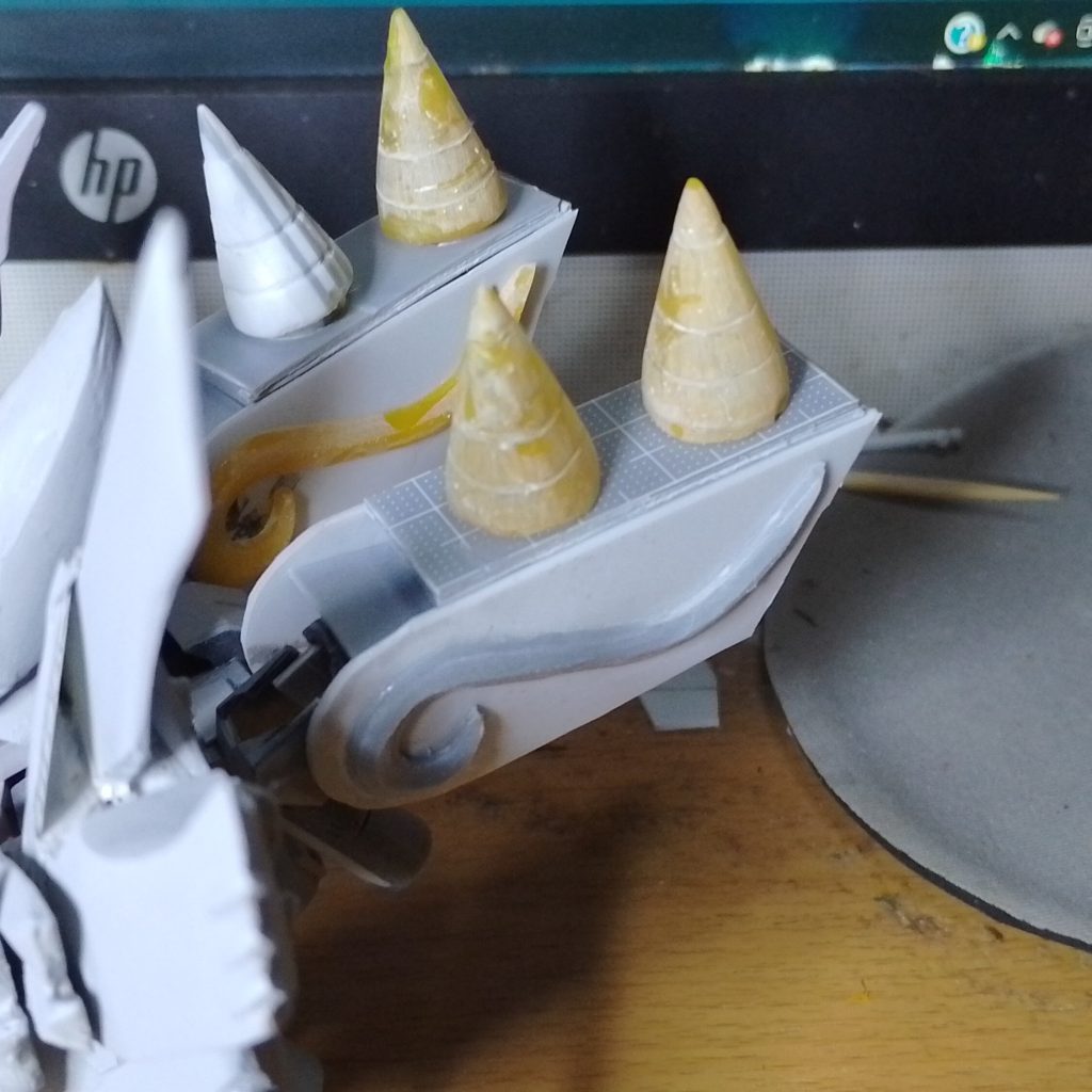 ファンネル(ミサイル?)プラ板を作った後に残った材料を電気コテで円錐形に成形し、彫刻線を研磨した後、UVパテで3枚にコピーしました。バックパック本体はオリジナルパーツを流用し、ファンネル基部を切断・再配置し、フィルムを貼り付けて大幅にサイズアップ。ベース両面のS字彫りはプラ板を大まかに切り出し、はみ出した端や角を電動グラインダーで切り出し、UVパテで3枚にコピーしました。