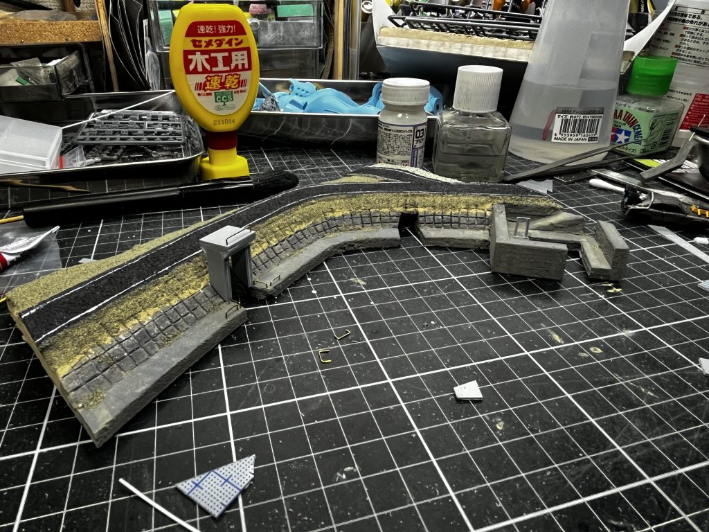 コルクシートを黒強めのグレー絵の具で塗って、ガイアのアルティメットホワイトで白線一発勝負。どなたかこれ以外の方法で細くて曲がった線を綺麗に引く方法あったら教えてください…ウェザリングペーストでコンクリート部分は下地を作り、質感を表現しました。その後ウェザリングカラーのマルチグレーて下地のサンドカラーが透けて見えるくらいのイメージで塗りました。ボンドベタベタに塗ってからジオラマ用の草の粉をパラパラしたらこんな感じです。雰囲気出てきましたね。あ、水門はプラ板積層とかでスクラッチしました。いろんな素材使うより結局プラ板が一番扱いやすい説、結構濃厚だと思います。