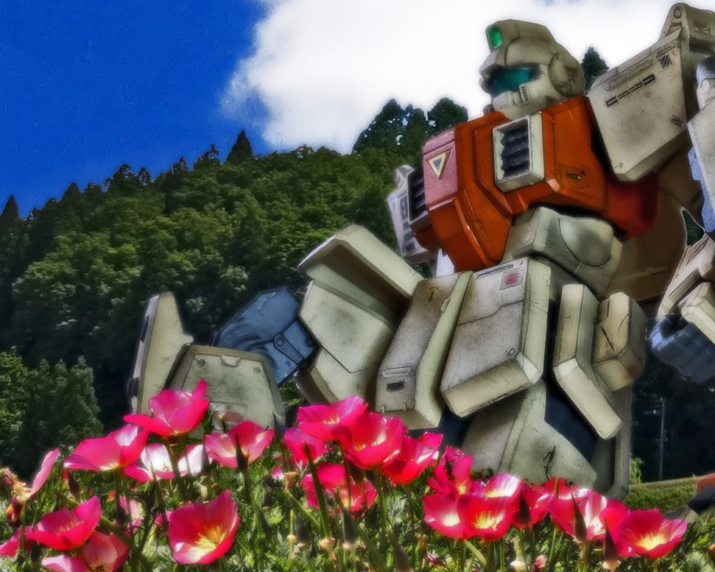 すると花畑の奥から濃紺のロングスカート姿の婦人が歩いてくる。「貴方達、連邦の兵隊さんがお菓子をくれるそうよ。もらってらっしゃい」「は〜い！」婦人は子供達をネスカから遠ざけるように軽く背中を押す。そのまま子供達は元気よく返事をすると膝をついたMSまで走っていった。「貴方は?」「ネスカ・コールマンです。サイド6のジャーナリストです」「あの子達に何か言いましたか?」婦人の顔つきは固く明らかにネスカを警戒している。「いえ何も。ここの花畑はどういったものなんですか?」「と言いますと?」「ここの植物は全部医薬品に使われるものばかりです…」婦人は花畑に視線をやると少し間を開けて話す。「ここでは地球連邦軍に卸す為の植物を栽培しているのです」「ではここは連邦政府の認可を受けて医療用植物の栽培をしているんですか?」「…そこは何とも言えないわ」「何故？」「連邦政府でなく連邦軍から委託されているのです」ネスカは婦人の言っている意味の理解に悩んだ。婦人は言葉を続ける。「村も若い人が減って、あのような子達にも大人の都合で始めた戦争の手伝いをさせているのは心苦しいものです。ですがこんな時だからこそ「命を育む」のを教えるのに植物の水やりをしてもらっているの」「情操教育の一環ですか?」「あの子達は嫌がらず進んでやってくれてます。この花が作られる背景は関係なく、何かできる事をしている事で子供達の精神的安定にも繋がっているのです」ネスカは複雑な心境であった。子供達が植物を育てるのは良いが、それに託けて間接的に戦争へ巻き込んでいるのではないだろうか。婦人は花畑を見渡す。 「破壊の中での創造…とでもいいましょうか」