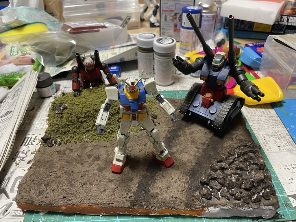100均でかってきた発泡スチロールに紙粘土と水溶きボンドで下地をつくって塗装。いまいちだったので、タミヤのテクスチャーペイントを塗ったら一気に土感がでました。あとは百均でかってきたジオラマ用の草とか石とかおいて固めました。石のところにタミヤのテクスチャーペイントのグレーを塗ったらアスファルトみたいになってしまったので、クレオスのウェザリングカラーでなんとかリカバー。ついでに思いつきでガンタンクのキャタピラ跡も塗ってみたら良い感じに。ガンダムはガンダムマーカー。タンクはランナー塗装。キャノンはエコプラにガンダムマーカーエアブラシ塗装。それぞれテーマを決めてやってみました。