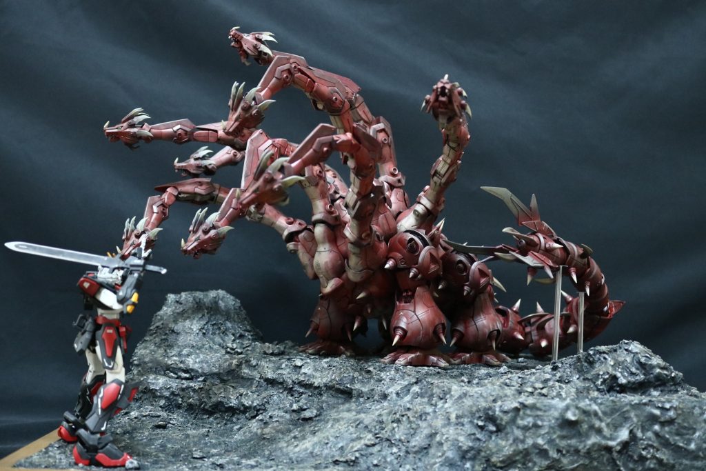 ヤマタノオロチとスサノオノミコトの対峙ヤマタノオロチが見下ろす。スサノオノミコトが剣を振り上げる。全体の配色を考えると、多色を採用すると情報が多すぎてしまうので、ジオラマのベースはグレー、ヤマタノオロチは赤、スサノヲノミコトは赤、黒、白を採用して落ち着いた雰囲気にまとめています。