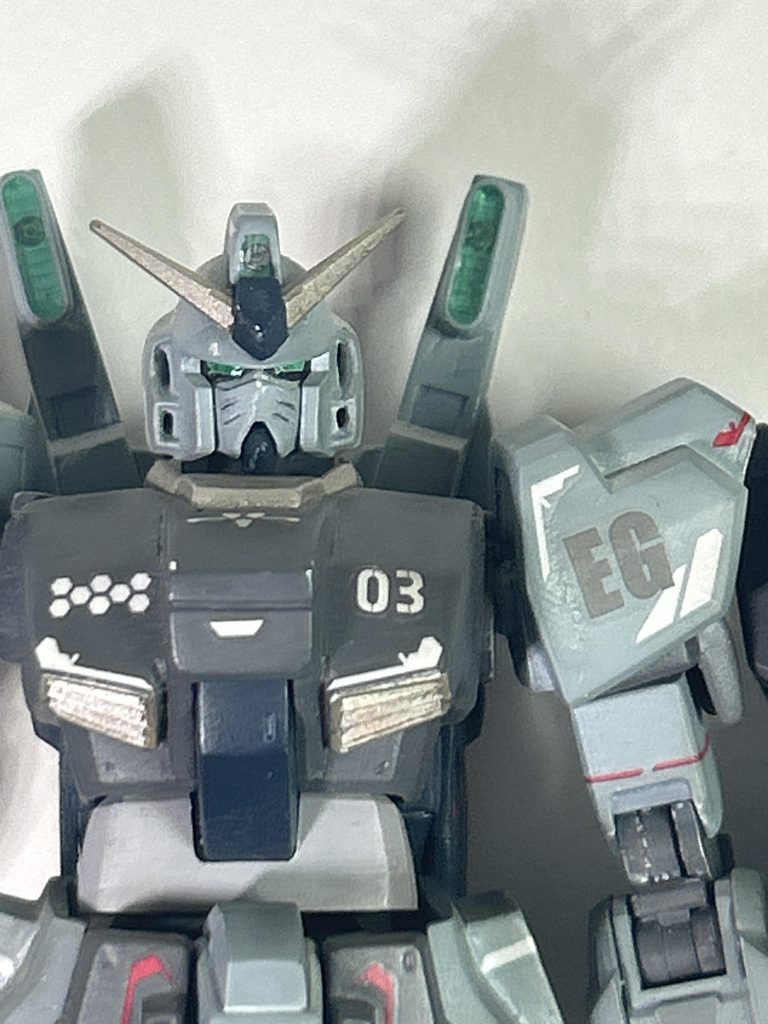 バストアップ。チャームポイントはマスクの猫ヒゲとインテーク設置位置変更の為に大きくなったバストサイズ。頭の加工部は前述通りマスク部のへの字を消して猫ヒゲ、メインカメラの大型化、頬のダクト穴の形状変更。胸部はカメラの切除、肩に繋がるフレーム上部を切断、胸をパテで盛って形状変更、コクピットブロックをプラ板で少し被せ気味に加工、腹部の延長。肩はノーマルのままで。バックパックは、ビームサーベルのラックを排除後にセンサー風な装備を設置、ブースター側面にプラ板にて整流板風に装着。センサー部は実はアポジモーターっぽいのではと気がつくのは後々のお話。なお、メインカメラとバックパックのセンサーはメタルパーツを埋め込みクリアレジンを流し込み作りました。