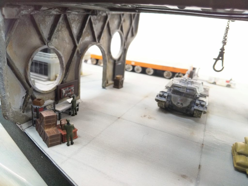 左の机、棚、木箱や、右に見切れている麻袋、後述するトラクター等は3Dプリント品を購入。わき役黒ちゃん(黒木製作所)Outland ModelsRS猿島地面＆格納庫の壁は、テクスチャーペイントの「ライトグレー」。これを塗るだけでリアルなコンクリート感。コテ・ヘラ状の板(プラ板でOK)で平らに均して塗った時と、厚塗りして乾燥後にヤスった時とで表情が変わるのも小ネタ。