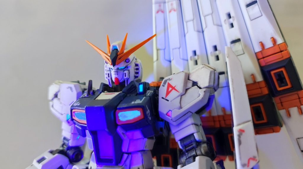 このビコーン！がやりたかった…。本当は本体の塗装は黄色部分のみ（ちょっと趣味に合わなかったので、オレンジ系を調色）としたかったのですが、白のプラの一部（肩の先端や腹部）も紫外線に反応してしまうため、やむなく艶消しホワイトを塗っています。また、ちょっと福岡版を意識し、左右肩にポジションライトの赤、緑を入れています。正規の位置はダクトっぽい意匠であったため、これは残すことにし、肩の下のほうの謎の台形部分をライトとしています。あまり大きな声では言えませんが、ホイル折り紙の表面に蛍光クリアを塗布し、適切な大きさにカットしてパーツの間に挟み込む、という荒業で作っています。パーツ分割の多いRGだからできた芸当です。