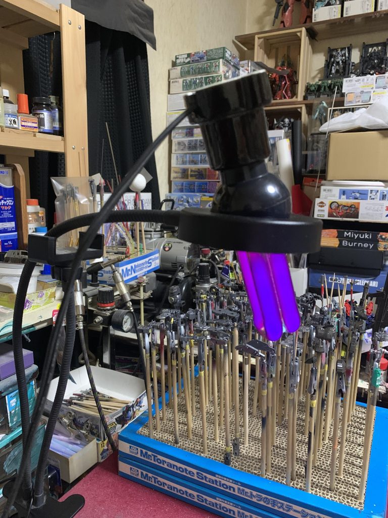 クワトロさんを塗る際に必須のアイテム。ブラックライト塗料はブラックライトを当てない限り透明なので、そのまま筆塗りしようとしてもどこに塗ってるかさっぱりわかりません。なのでブラックライトを照射しながらの作業になります。ブラックライトは机に置いて、手元を見えやすくする為にリングライト(通常光)でクワトロさんを照らすとブラックライト反応が弱まるので、やっぱり見えません。そこで、リングライトの穴にブラックライトを突っ込んで、部屋の照明は消してリングライトの明るさを弱めつつブラックライト照射でやっと作業が可能になりました。ブラックライトの角度と距離、リングライトの明るさのバランスが最適じゃないと不可能に近いです。塗りの技術より、いかに見えるようにするかが大事です(笑)