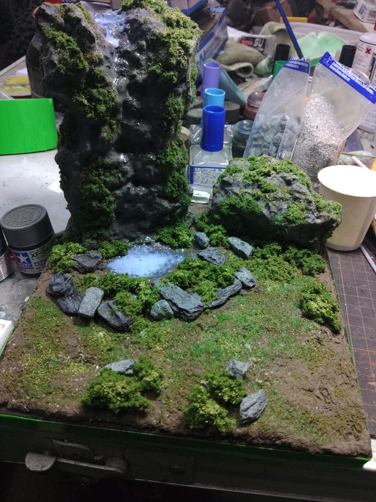 ジオラマ部分はストックしてあった素材や100均やホームセンターの商品を使用して、なるべく低コストで作りました。岸壁や大きな岩は、発泡スチロールに、100均の粘土を木工ボンド、水、グレーの塗料で溶いた物を塗りつけています。小さな岩は市販の石素材と、バークチップに着色した物を使用しました。草等はストックしてあった素材を適当に盛り付けています。滝壺は、青で着色した上に、グロスポリマーメディウム、ジェルメディウムを盛っています。