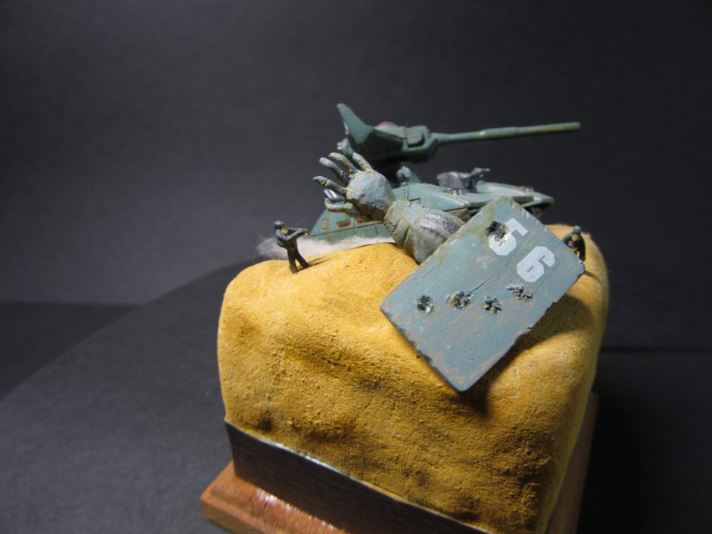 ザクの腕。キットの腕は握り手なので指部を切り落とし、アルミ線で開いた指を表現。敗れたことに対する無念さを表現してみました。またシールドはプラ板にて大型化。砲撃によるダメージ表現も追加しています。ベースはスチレンボードを切り出し、積み重ね木工用ボンドで固定。上から木粉粘土で砂漠地帯を表現。表面を手ぬぐいで包んで細かな表面地にしてみました。外観は少し斜めに仕上げマゼラアタックに動きが付くようにしています。また側面には木目調テープでデコレーション。