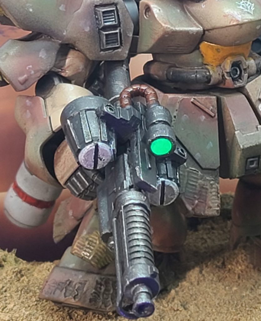 一方、こちらのマシンガンのセンサー部分はいつものように蛍光塗料ビコーンシステムです(笑)。いつもとちがうのは裏をシルバーではなくホワイトに塗っているということ。その方がブラックライトなど当てなくても発色がよく、光っているようにも見えるようです。マシンガン本体は、ビルダーズパーツジオン系武器セットのザクワンマシンガンをベースにでっちあげました(笑)特徴的な左右のドラムマガジン?は市販のバーニアパーツをベースに作成。バレルの形状などはイラストと違いますが、バレル下のガードフレームや左右のドラムマガジン、上部のセンサー、それに繋がっている動力パイプみたいケーブル、という記号が揃えばそれっぽく見えるかなぁと。