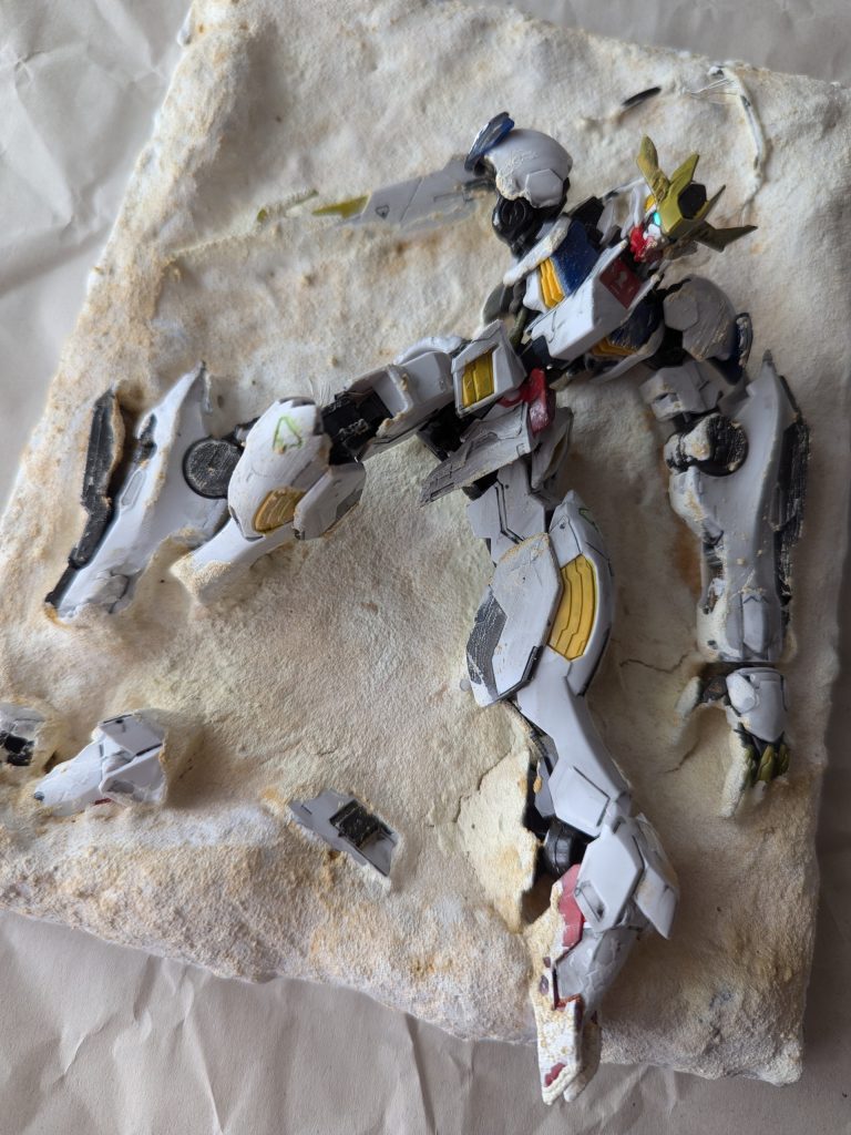 100円の写真立てをベースに同じく100円の粘土で土台を作り、バルバトスルプスレクスを埋める位置を決めて印をつけて乾燥。乾燥後にルプスレクスを置いて接着剤で接着→乾燥。乾燥後に再び粘土で接着した部分を消しつつルプスレクスを埋めていきます。土台の一部には子どもが遊んだ発掘して遊ぶおもちゃの余った砂を使っています。