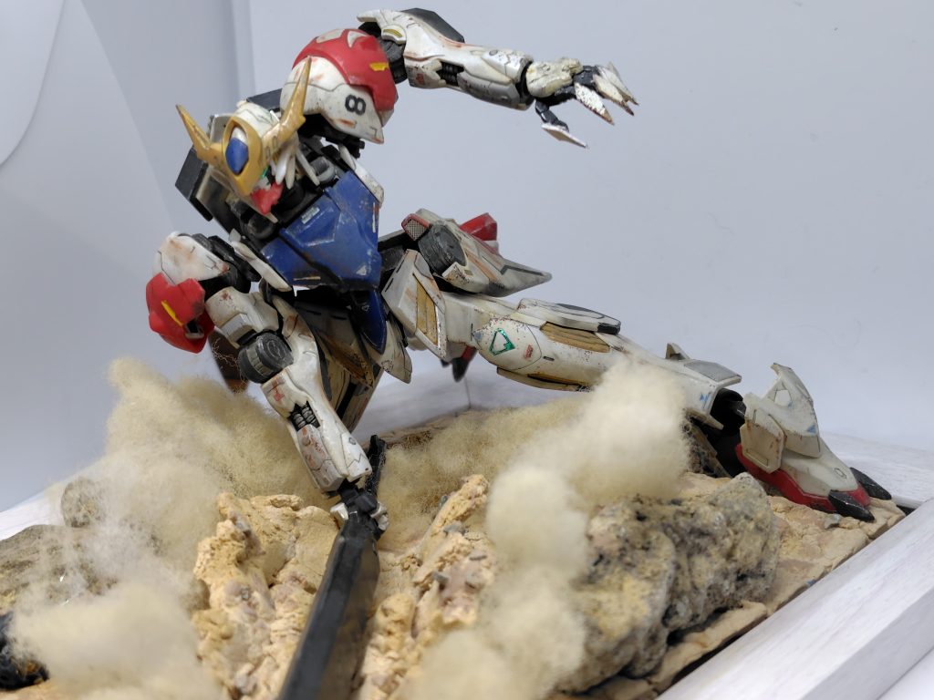 ジオラマ部分 砂地でメイスを叩きつけるバルバトスをイメージしました。外側の枠は100均の写真を飾る額縁です。地面の部分は紙粘土で作り、そこに石をおいてウェザリングしました。全体的に園芸用の小石を散りばめ、綿で土煙を再現しました。