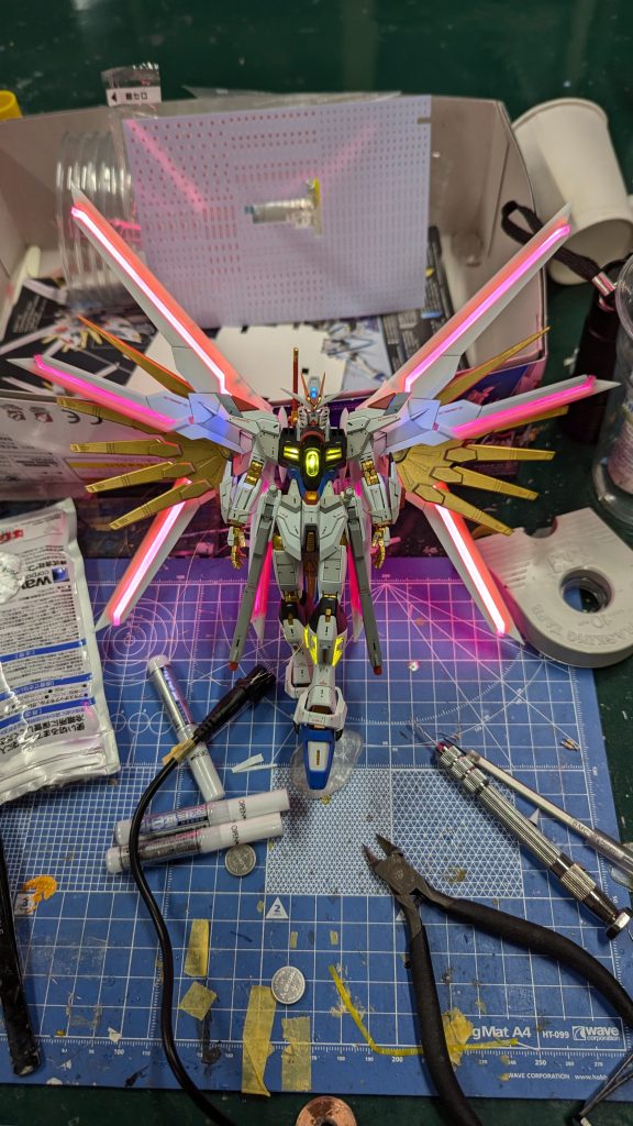 電飾が終わった状態印象的なピンク発光部分はピンク発光のフィラメントLED厚手のクリアファイルを使って再現しています。本体の電飾は全て1608白色LEDで行っており、色はその上に被せてある着色したUVレジンによって着けられています。導線は胴体部に集まっていて、股の3ミリ穴から出てくるようにしています。特に大変だったのは頭の電飾です。クリアパーツが青のせいで目まで青になってしまうという問題があったのですが、LEDとクリアパーツ表面間の空間を黄色のUVレジンで埋めることで目を黄色に光らせることが出来ました。