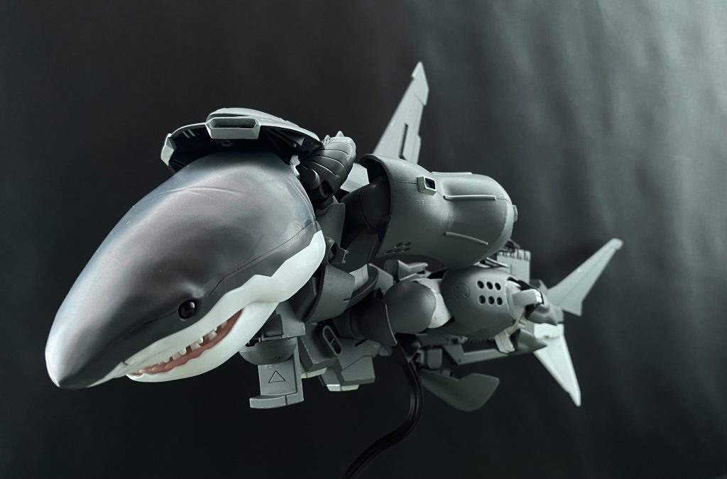 　海上の光も届かないほど深い、暗い海の中を巡航形態となったシャークゴッグが泳ぐ。MS状態における背部から腰部にかけて長距離航行ユニットを装着されたシャークゴッグは、その名のとおり巨大な「サメ」のシルエットを暗い深海に浮かび上がらせている。…20メートルをゆうに超えるその姿は、太古の昔、この海に王者として君臨したメガロドンを彷彿とさせる。差し詰め、「メカメガロドン」と、いったところか。　…いや、まて。僕は何を言っている？落ち着け。オリヴァー･マイ。…深呼吸だ。…よし。　気を取り直し、改めてモニターの向こうにいる、シャークゴッグのパイロットに声をかける。「ワシヤ中尉、こちらからの機体チェックはオールグリーン。中尉の方でよろしければ、評価試験を開始してください」　今回、この機体のパイロットとして抜擢されたヒデト･ワシヤ中尉だったが、モニターの向こうの中尉の表情は暗い。それもそのはず。彼の乗っているシャークゴッグは、前回のテスト時に暴走している。いや、正確には彼の機体は二番機であり、暴走した機体ではなく新たに組み上げられた完全に新規の機体だ。当然、暴走の原因だと思われたOSも別のものに変更されている。「えぇ〜、二番機、ですか？」「あら、ヅダの時は喜んでいたでしょう？」「…うむ、海の男である中尉にしか頼めないことだ。すまないな」　出撃前の艦長らの会話を思い出す。大丈夫ですよ中尉。と声をかけて見たものの、正直なところ自分もこの機体に対してあまり良い印象を持ってはいないのだ。それはワシヤ中尉も同じなのだと思う。「…コレ、本当に暴走しませんよね」　深海にワシヤ中尉の声が響く。そうして、MSM-03Sシャークゴッグの評価試験が開始された。