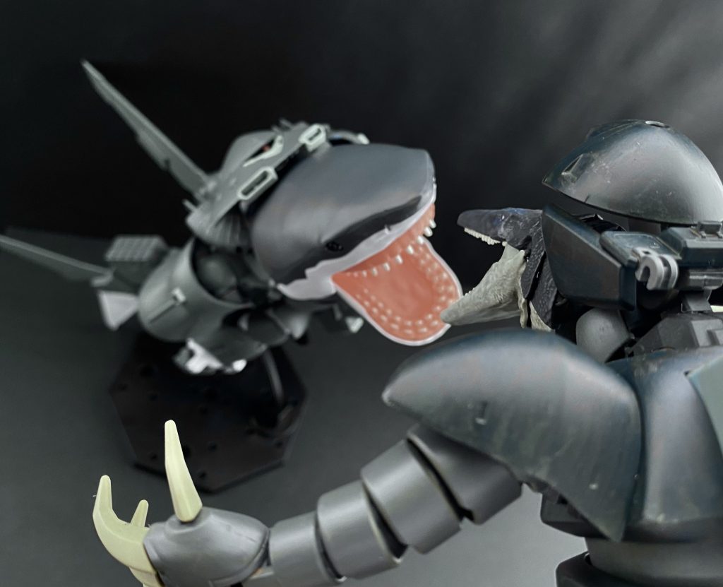 　水中での機動戦闘。互いに激しく動き回る為、魚雷を始めとした火器の使用はあまり有効ではない。その為か、水中での運用を考えられたMS、MAには大型のクロー状の武装が装備され、接近戦闘を見据えた装備構成になっていることが多い。それは中尉の乗るシャークゴッグも同じだ。しかし、今、シャークゴッグは長距離航行する為のユニットを装備している。この機体ベースとなったハイゴッグにおける巡航形態の状態でユニットを装着することで、長距離の高速、静音航行を可能にしているのだ。そのかわり、MSの腕部として折りたたまれている腕部クローを展開出来ない。…つまり、今、あのシャークゴッグに残された近接用の武装は、あの大きな顎しかないのだ。シャークゴッグはその名の通り「魚類」として戦うほか、ないのだ。　僕達、宇宙で生まれ育ったスペースノイドは知識として海を、魚類を知っていても、本物の海を、そして「サメ」を知らない。…それは今、パイロットとしてあの機体を「操縦」しているワシヤ中尉も同じなのだ。…モニターの中に映るシャークゴッグの動きを通して、中尉の焦りが伝わってくる。　「落ち着いて、中尉。…まずは相手との距離離しましょう！」　そう言ってはみたものの機動性能は明らかに相手の方が上だ。「無茶だっ、あっちの方が素早いっ！…そうだっ、航行用ユニットをパージすればっ」「だめですっ、中尉！…そのユニットをパージすると、中尉が帰って来れません！この大海原で、溺れてしまいますっ」　そう、あのユニットはあくまで機体の長距離航行用のユニット。機体への着脱は専用の施設がなくては出来ない。たしかに、ユニットをパージし、シャークゴッグをMSとして戦闘機動させれば、相手に対して互角、それ以上の戦いが出来るだろう。しかし、それではワシヤ中尉が帰って来られないのだ。　モニターの中では、まるで生き物のように動くゴッグ？と、サメのように動くゴッグがぶつかり合う。顎と爪が、顎と顎が。そうして何度目かの接近の時、モニターの中のワシヤ中尉が叫ぶ。「…嘘でしょっ！直上っ、熱源反応っ！」「中尉っ！回避をっ！」　それと同時に、二機の間を裂くように戦艦の主砲のようなビームが放たれる。「そんなっ、ビーム砲、水中であれほど強力な…」　モニターを見ると、ワシヤ中尉は無事、回避したようだ。そして相手のゴッグ？も。しかし、上を見上げたシャークゴッグのメインカメラからの映像を見て、その場にいた者全員が凍りつく。遥か海面からの光を背に、人型のシルエットが浮かび上がる。「あ、あれは…、ガンダム」　RX-78ガンダム。連邦軍が開発したMS。そのMSはまるで人間のようにくるりと頭を下に回転すると、手足で大きく水を掻き、泳ぎ、こちらに向かって潜ってくる。…そして、驚く僕達を後目に言う。…オープンチャンネルで。「オイオイ、本気(マジ)か。…巨大な(デカい)サメに、巨大な(デカい)ワニか？…昨日の酒はもう抜けたと思ってたんだがな」