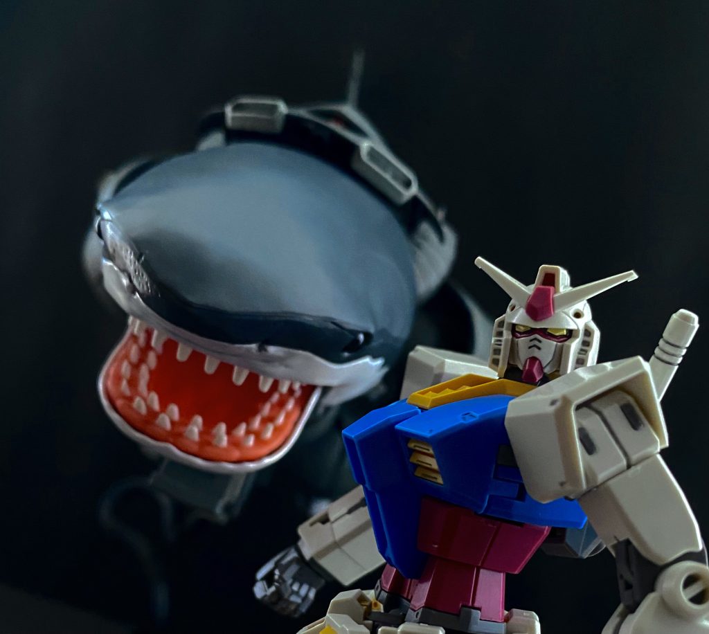 「おいっ！聞こえるか、サメ野郎っ」　シャークゴッグのコクピット内に男の声が響く。いきなりオープン回線での通信に動揺を隠せないワシヤ中尉は、思わず返答してしまう。「えぇ、オープンチャンネル…、お、オレはジオンだそっ！」「ああ？ジオンだろうがなんだろうが関係ねぇ。あっちのワニ頭は、返答も無いからな」　…普通は、返答しないと思うのだが。そんなツッコミをすんでの所で飲み込んだワシヤ中尉は、目の前の状況に警戒しながら、この状況を記録しているだろうオリヴァー･マイへの通信をしようとするが、繋がらない。どうやら離れ過ぎたらしい。「おまえらはなんだ？何で身内でやり合ってる？…脱走兵か？」　矢継ぎ早に話かけてくる連邦のパイロットの声を聞きながらワシヤ中尉は考える。今、目の前に敵がいる、連邦軍。しかも、白い悪魔と呼ばれ恐れられているRX-78ガンダムが。そこまではわかる。解せないのは、攻撃の意思がない。…のか。「お前らは、ここから先に用があるわけじゃ無いんだな」「そ、そうだ。オレ達は試験中に襲われただけだ。…この先に何が、あっ」　簡易マップを開き示された位置を確認する。そこには、大規模な海上油田プラント群が。…気が付かなかった。「いいか、サメ野郎。俺達はお前らにあそこに行って欲しくない。…お前はどうだ？このままならあのワニ頭は、真っ直ぐプラントに行きかねん。どうする！サメ野郎！」「サメ野郎って言うな！オレはジオン公…」「おっと、自己紹介はいい…。次に会う時は敵だからな。…よし、なら共闘だ。やるぞサメ野郎！」　共闘って何を言い出すんだこの連邦兵…。コクピットの中、ひとり頭を抱えるワシヤ中尉だったが、技術中尉らのユーコン級と逸れてしまった今、二機を相手に立ち回るののは得策では無い。あのゴッグ?だけでもかなり相手だ。それと同時にガンダムを相手にするなどもってのほか。ならば、こちらに攻撃の意思がないガンダムと、一時的とはいえ共闘した方が生き残る確率は上がるだろう。それに、このシャークゴッグの他にガンダムのデータも取得出来るかもしれない。後の事はユーコン級と合流してから考えればいい。「わ、わかった…」「よし…、それでいい。お前の機動力を貸せ！サメ野郎、…おかしなマネしたら後ろから撃つからな」「えぇ…」