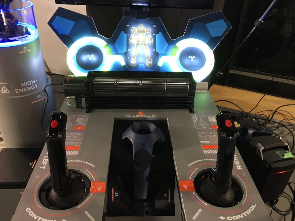 赤いボタンとトリガーは全て攻撃コマンドなのでどれ押してもOKな仕様です。 筐体に座るとこんな感じ。後述しますがVRヘッドセットを付けると左右レバーの操作でどう移動するか説明が出てきます。真ん中にあるのは出撃前に3Dデータ化したガンプラを手に取って見られるコントローラーです。