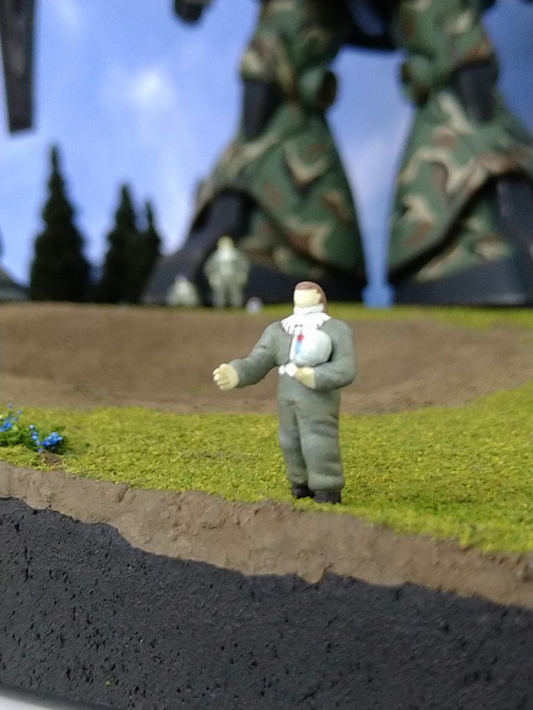 主役である隊長パイロットのアップ。本作のきっかけは、去年の4月、過去作の『MS-09 破片迷彩』を作った事でした。そこから3機小隊でのジオラマを作りたい気持ちになり、・場所はオデッサ(現ウクライナ)辺り・ザク、グフ、ドムの3機を使用といった辺りはスッと決まったものの、『どんなシーンにしようかな』と素材を探す内に行き詰まり、連日報じられるウクライナの現状を見るにつけ、何となく今回は『シンプルに、敵味方で握手をする画を見たい』という思いが強くなりました。(★★特定の思想や政治的意図はありません★★)で、その画を頭に浮かべると設定や作業も一気に進み出しました。特に写真の人物作成において、握手を求める右手はこの作品最大の『肝』であり、一番注力した箇所ですが、『優しく、柔らかく、ややすくい上げる様な』感じが出た気がしました。1/100でここまでやれたなら、自分としては大満足で『御の字』です😆😆😆★さて、ここで以下に作品の基礎設定を記します。作品の味わいが少し増すかもしれませんので、お手すきな方はご覧下されば幸いです。(勿論スッ飛ばして頂いて結構です😊)≪ ジオラマ向け設定(妄想) ≫【基礎設定】・3機は、マ・クベ麾下の虎の子部隊のひとつ、『第1特別MS大隊』の第4中隊に所属。・第1特別MS大隊の構成は4個中隊からなる。通常の機械化大隊は3個中隊で構成されるが、本大隊には特別に第4中隊として、先行攪乱や陽動、威力偵察を行う中隊を保有。第4中隊による攪乱後、第1~3中隊で一気に敵陣を殲滅する意図。・上述の理由により、第4中隊は熟練パイロットで固められていた。・第4中隊は象徴的な精鋭部隊である為、中隊規模では異例の部隊章を認められており、『デスサイズ(死神の鎌)』を制定している。・また第4中隊は難易度の高い任務を行う為、敵中で瞬時に味方の識別を行うべく、独自の部隊カラー(マルーン)を機体の一部に塗布、同士討ち回避の対策としている。・機体番号は3桁で各桁の説明は以下。 1桁目∶所属中隊番号。 2桁目∶所属小隊番号。中隊本部は0。 3桁目∶小隊内機体番号。隊長機は0。【場所及び時期】 オデッサ近郊、0079晩秋〜0080初夏。【背景】 要衝オデッサは陥落し、第1特別MS大隊も壊滅的被害を受けたものの、第4中隊は16機の内12機が残存。(中隊本部は4機の内、1号機を損失。) マ・クベは予め、万一のオデッサ失陥の対応策として、秘匿した小規模地下拠点を多数準備しており、連邦占領地域でのゲリラ攪乱を仕掛け続け、連邦戦力の宇宙への集中を遅延させる為の戦力を残していた。(その戦力の宇宙脱出拠点も一応準備済。) 殲滅を逃れた第4中隊もこの戦力に合流、小規模拠点ごとにMSを分散して配置、継続した抵抗を続けていた。そこに支援組織も現れ、終戦後も抵抗は続く。しかしやがて。。。