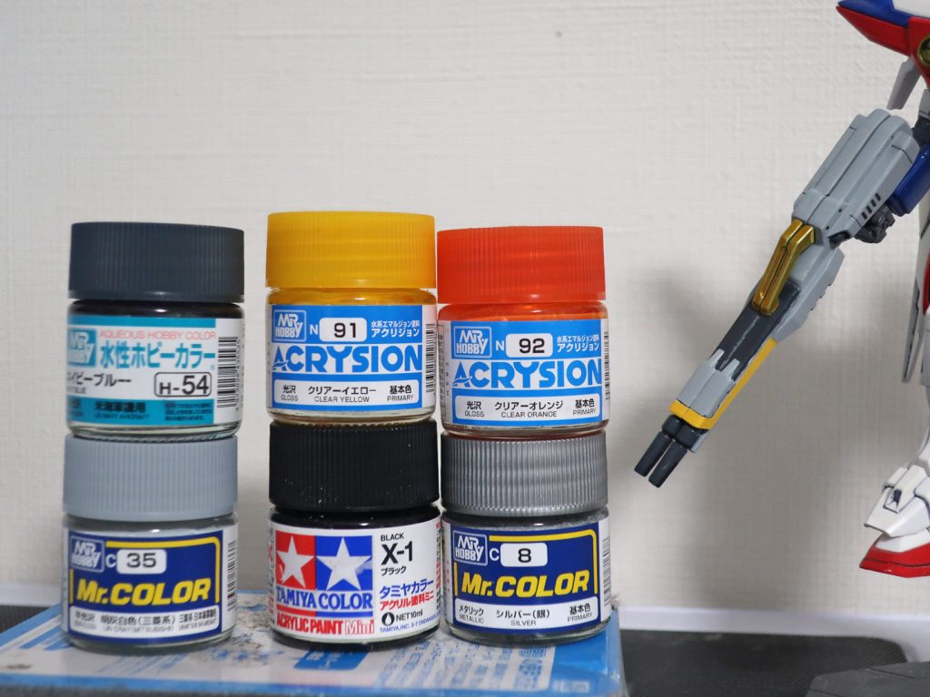地味に一番考える時間が長かったライフルのメイン色。W公式ページの設定画だと銃身は薄紫系のグレーなのですがアニメ本編では白寄りのグレー（ライトグレー）だったのでヨドバシの塗料売り場で延々と塗料とにらめっこした結果写真の左下の明灰白色を選びました。自分の中でこれは完璧とはいかないまでも8割くらいは満足できたのでまあいいのかなという感じです。逆に銃口やグリップはネイビーブルーを使用しましたがこちらはやや暗かったかもしれません。ホワイトを微量混ぜるのもアリかって気もしたのですが作業量を考えて単色で仕上げました。今になって思えばニュートラルグレーが一番かなという気もします。上部のゴールド部分はブラック下地でシルバーの上にクリアーイエロー+クリアーオレンジ少量という定番レシでその上に半光沢トップコート。当初はラッカー系で筆塗り3段で挑みましたがさすがに何度も失敗したので最終的に一番上層のクリア部分はアクリジョンに頼る事でなんとか切り抜けました。薄く吹けるエアブラシがあれば…( ´；ω；｀)上の写真ではブラックがタミヤカラーのアクリルになっていますがこれは本来望ましくない組み合わせなので最下層はラッカーのブラックが良いと思います。（幸い今回はその上のシルバーはキレイに発色してくれましたが）
