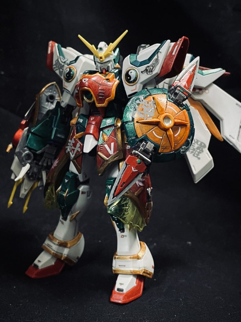 シェンロンガンダムはなんか地味な印象でした。なので、武神のイメージで哪吒っぽくしていこうと改修を始めました。弁慶のように多くの武器を携行するようにした為、シェンロン自体が華奢な印象になりましたので、横に大きくするように意識しました。肩はジャンクの足パーツを使って肩幅を大きく胸は元々開きそうなアーマー形状だったので、カットしてからずらしてパーツ追加して、ウイングガンダムのような形にしました。腰は和風武器のパーツを使って胸パーツの様に、緑の装甲に鎧をまとっている印象になる様意識しました。BBからクリアパーツを拝借し、全く効果のない装飾として取り付け。　足自体は色付けぐらいしかやっていません。バックパックはハルートのバックパックと、フーティエを連想できるベギルベウのバックパックをミキシングして作成。腕の伸びる機能はオミットして、手甲と、ガトリングに「シェンロン＝龍」の要素を残しました。「ナタク＝哪吒」として、近接特化としましたが、哪吒の話には遠距離武装と思われるものもありましたので、バックパックにガトリングを装備させました。