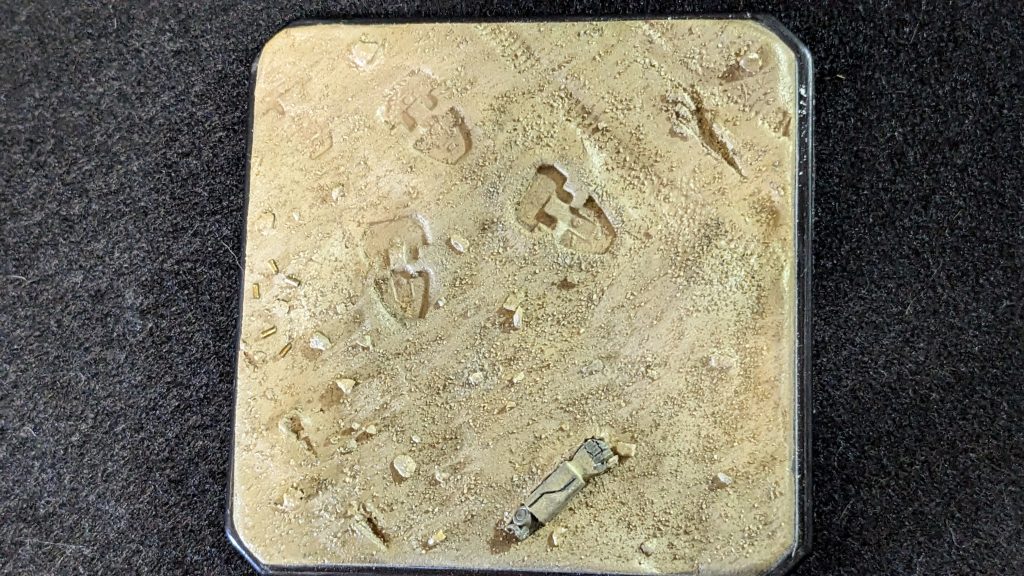 100均で買って来たケースを使いました。もう少し小さい物はないかと探していたのですが、思った様なサイズの物は無く(有ったには有ったのですが、蓋が無かった・・・)仕方なしです。とか思ってたのですが、いざ完成させると割りと良いサイズなのではと思ってますwただこのサイズなら、もう少しストラクチャーを作れば良かったかな。それこそ墜落したフライマンタとか。ダイソーのディスプレイケースにセリアの造形粘土で作ってます。少し起伏を作ったベースにプラ角材で作ったGMの腕を配置。ザクの足跡とWTMの戦車を使ってキャタピラの跡をスタンプ。水で溶いた木工ボンドをザクとGMの腕を固定する穴以外の部分に塗って本物の小石をふりかけ、本物の砂を篩を使って振り掛けました。 最後に金色に塗った伸ばしランナーを短く切って足元に配置しました。飛ばした空薬莢のつもりです。