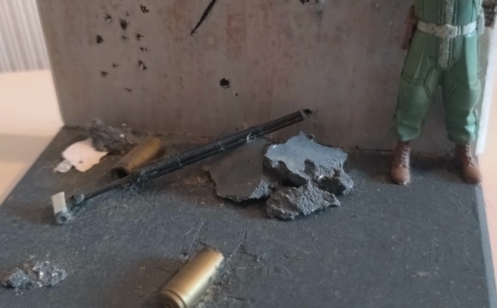 建物の損壊度の割に、地面が綺麗すぎますね。申し訳程度の瓦礫と、ザクマシンガンの薬莢を配置してます。真ん中の黒い棒は、ビデオカメラと三脚のつもりです。しょうもないですが、わざわざこのジオラマのためにスクラッチしました。当時はまだこの形式のビデオカメラは現役で使われていましたが、今ではすっかり見なくなりましたね。宇宙世紀ではとんでもない骨董品として扱われているんじゃないでしょうか。