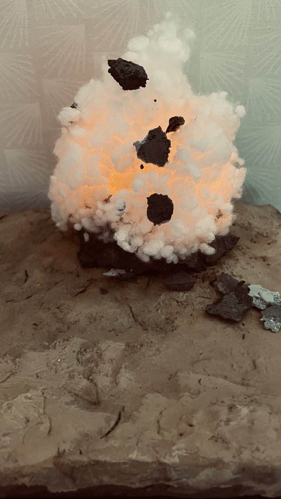 爆発エフェクトのアップです。100均のピンポン玉(オレンジ色)に同じく100均のLEDキャンドルを差し込み爆発の光を表現してみました。爆炎は手芸用つぶわたにアクリルの黒/オレンジを吹いています。(写真は着色前の状態) つぶわたの固定を意図して盛り付けた後木工用ボンドを水で溶いたものを上から吹き付けるとよいと思います。噴き上がる土のかけらは粘土をのして細かく砕いた物です。