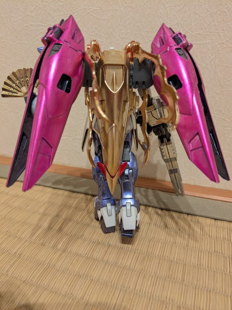 バックショットを1枚。ウイングは彼女の持つ傘をイメージしてメタリックピンクに塗装。シールドはバックパックに下向きに装備し、イエローゴールドで塗装し、彼女の長髪っぽく見えるようにしてみました。シールドの小羽根とカメラアイはロイヤルメタレッドに塗装し、髪を束ねたリボンに見立てています。挟み込んだ炎エフェクトが前だけでなく、後ろや下にも出ていて、躍動感がある感じになりました。