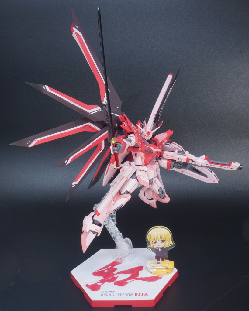 以下、細かな設定です。オーブにおいて実体剣「フツノミタマ」が開発されたが、オーブでは初の実体剣であったため、運用にあたり実証実験が必要になった。キラ准将が使用することを想定していたため、准将の搭乗機であるライジングフリーダムでの実証実験が適切と判断され、予備パーツによりライジングフリーダムの2番機が建造された。基本スペックは准将が搭乗したライジングフリーダムと同等であったが、フツノミタマの運用のために装甲強度を上げており、結果VPS装甲は赤系統の配色へ変化している。また、装甲強度に電力配分を多くしたために火器系統への電力配分が少なくなっており、火器の攻撃力は通常設定のライジングフリーダムより劣るが、機体を通常設定で再起動すればライジングフリーダムと同スペックで運用することは可能である。(この場合装甲強度も通常設定のライジングフリーダムと同等となり、機体色も青になる)フツノミタマは御神刀としての建造でもあったため、制式採用され准将が携行した「真打」以外にも数本の「影打」が存在し、実証実験においては「影打」が使用された。実証実験を終えた本機は再びライジングフリーダムの予備パーツへと解体される予定であったが、実証実験を視察したカガリ代表が自身の搭乗機であったストライクルージュと似た機体色であった本機を気に入り、「ライジングフリーダムルージュ」と名付けて代表専用機として配備されることとなった。ただ、代表が戦線に出ることを案じたオーブ側近たちが整備スタッフに搭載OSの最適化を命じ、「調整」と称して時間稼ぎすることにより本機の代表への配備を遅らせていたが、そのうちにファウンデーション事変が勃発。本機の配備が間に合わず最終的に代表は旧式のストライクルージュで戦線に出てしまったため、事変収束後、改めて本機の調整が行われ、正式に代表の下へ届けられる運びとなった。