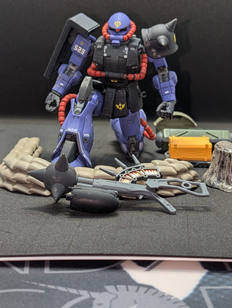 マレット「……っち、仕方ない片方だけは試してやる。で？コレはどういう兵装だ？」(でん、でででででっでん、でででででででっでん)マレットには、何かBGMが聞こえたような気がした。マイ「はい、説明します。コレは[特殊ハンマー・ガン試作型]、ペズン工廠において開発中の[MS-13 ガッシャ]の携行兵装として開発が進められているものです。見ての通り、巨大な鉄球を打ち出すもので、銃身下部にドラムリールを装着し、鉄球とは有線で接続され、発射後はドラムリールによって引き戻され再装填されます。銃身上部に装着されたマガジンの弾薬は120mmザクマシンガンと同じ物から弾頭を抜いた物を使用しており、1コッキングで3発が一度に薬室内に装填されます。3発を同時に撃発し、発生した燃焼ガスで鉄球を加速します。その分、発射時の反動はかなりのものとなりますので、折り畳み式ストックが装着されています。片腕で撃つのはザクの腕部には負荷が大きすぎると思われますので、必ず両腕で撃つようにして下さい」マレット「…ふむ。聞いているとこちらは少しマシなもののようだな。それに、臆病者の連邦軍にはこういった「痛そうな形」のもののほうが効きそうだ。」マイ「そういうものですか？」マレット「ああ。想像してみろ。パイロットは、コクピットの映像を通して自分の目で見ている。人間の争いが小さな銃弾の交わし合いになった現代で、この巨大な鉄球が自分を潰しにくるその様は、恐怖だろうな。」マイ「は……」マイは、マレットの目の奥の狂気を垣間見えた気がして、身震いをした。