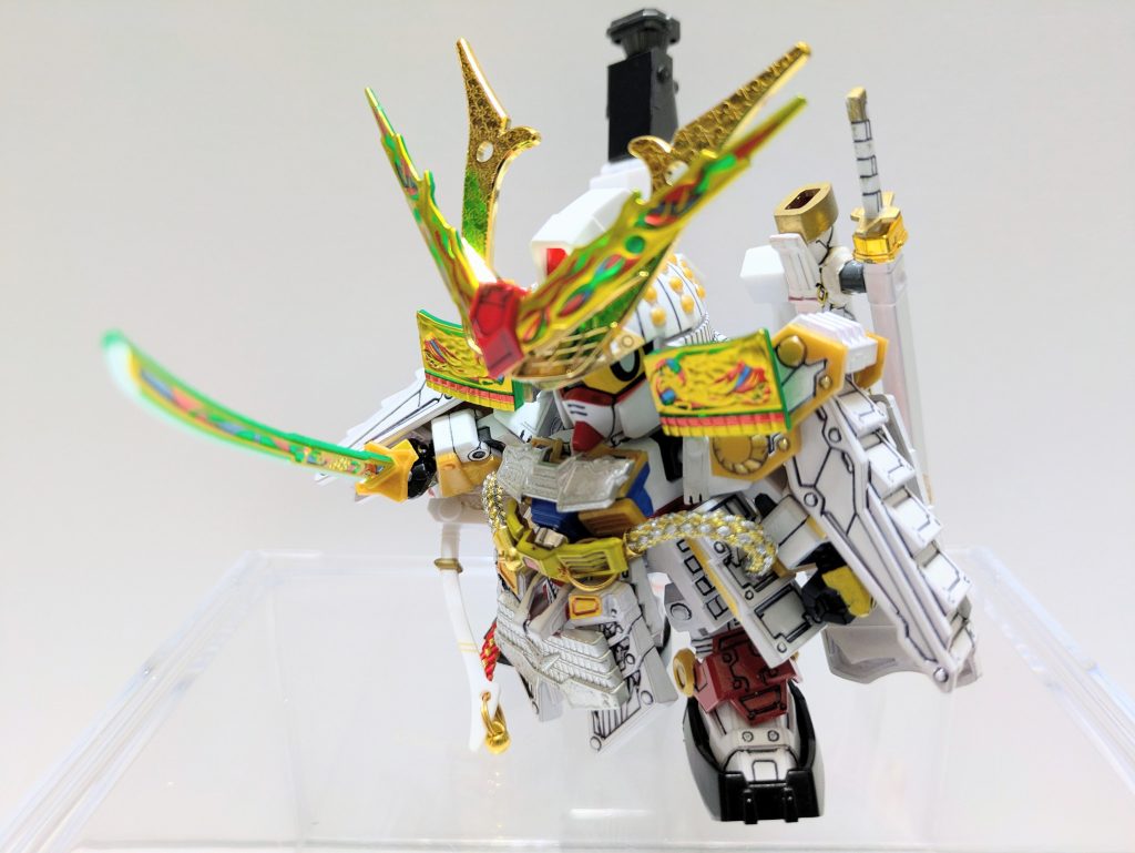 私説・SD戦国伝/弐十國戦記〜光と闇の大戦〜【第1話/混沌】遥か太古。母なる宇宙は創生した地球に生命を育むために全知全能の力を持たせた双子の神を生み落とした。一方には正義を、一方には野心を与えて。兄弟は力を合わせ、時に競い合いながら平和を築き上げていった。だが、時を重ねるにつれて両者の心に眠る相反する信念は軋轢を生み、やがて決定的に決裂する。伝承のみに残る〝光と闇の大戦〟によって両勢力はとうとう武力によって衝突。気の遠くなるような時間と多くの犠牲を払いながらも雌雄は決さず。激怒した母なる宇宙は地球を二つに分け、未来永劫互いが干渉しないよう地上には結界を張り、魔界には呪縛を巡らせた。そして、兄弟の余りある力を〝光と闇の神器〟に吸い上げ、各界に散らした。力を失った光の神は「天聖大光帝」を名乗り地上を統治、闇の神は「闇黑魔大帝」として魔界へと消えた。時は流れ、泰平の世。神々は伝説となり、戦乱は夢物語となった世界。光の神器を国宝として祀る地上の弐十國の一つ、天宮の皇子・白帝頑駄無は民からの信頼を厚く武芸百般に秀でる最強の武者として日々の鍛錬や武士育成に精を出していた。だが、それはあくまで防衛や腕を高め合うためであり、誰一人自らが実戦に身を投じる未来が現実に訪れようとは夢にも思わず。そんなある日、突如として結界に衝撃が走った。闇の気が流れ込み、各地で天変地異を引き起こす。各国を統治する大将軍たちが次々と奇病に倒れていき、地上は混沌に包まれていった。神の力以外では絶対に破られない結界にいったい何が起こったのか。「白帝よ…。永らく眠っていた闇の神が力を取り戻しつつあるのかもしれぬ。一刻も早く結界を修復し、闇の気を絶たねば血で血を洗う戦いが始まることになる。魔界の門に向かい、異変の正体を突き止めてくれ」病床の父から光の神器〝武輝丸〟を託された白帝は異変の解明と平和を取り戻すために幼なじみの頑巨砲、鉄機武者の頑戦車と共に旅に出る。果たして彼らを待ち受けるものとは…。次回を待て!!